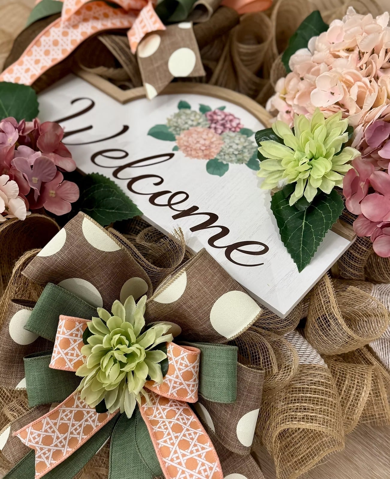 A decorative rustic wreath with a white heart-shaped 'Welcome' sign, faux flowers in pink, peach, and green, burlap ribbon, and patterned bows in brown, peach, and sage green.